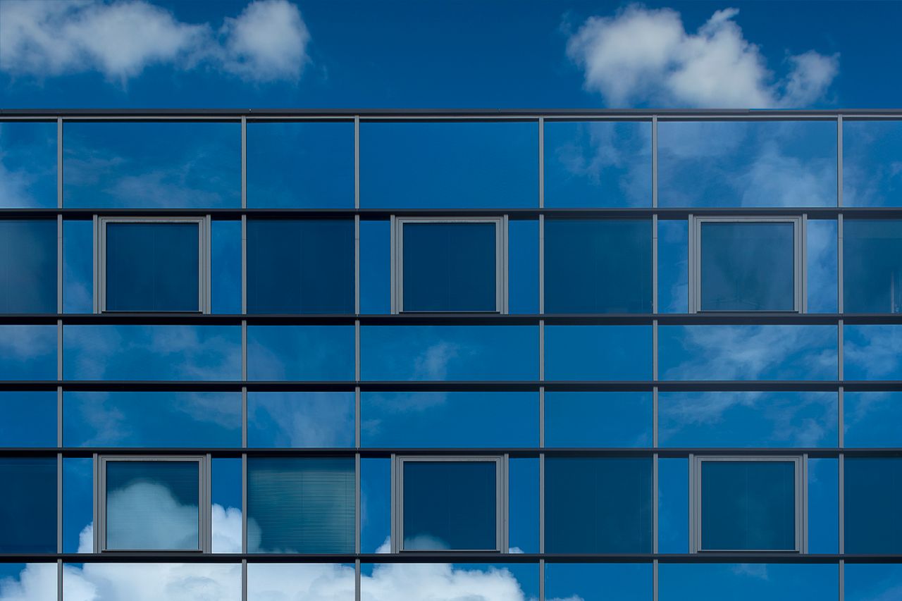 hero-img-01 Facade of a modern glass building reflecting the blue sky and clouds.